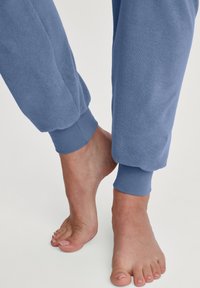 Hellblaue Sweatpants mit gerippten Bündchen an den Knöcheln, die eine weiche Textur zeigen. Die Füße sind barfuß, mit manikürten Zehen, in einer neutralen Umgebung positioniert.