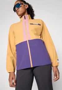 Žena koja nosi naočale za sunce i zip-up jaknu u boji žute, ružičaste i ljubičaste s crnim hlačama na jednobrojnoj pozadini.