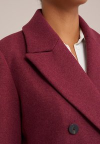 Burgundy gyapjú keverék blézer, kivágott gallérral, dupla gombolású kialakítással és egy fekete gombbal. Texturált szövet részlet látható.