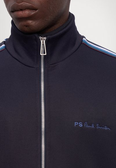 Marinblå zip-up träningsjacka med hög krage, med en silverdragkedja, broderad "PS Paul Smith" logotyp och flerfärgade ränder längs axlarna.