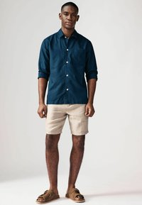 Camisa de lino azul oscuro con mangas arremangadas, bolsillo en el pecho, combinada con pantalones cortos beige claros. El modelo lleva sandalias marrones.