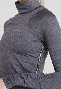 Haut de sport à manches longues gris avec un col montant, doté d'un panneau latéral noir à logo et d'une coupe ajustée. Confectionné dans un tissu lisse et extensible.