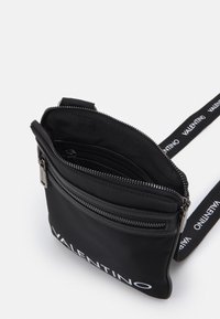 Sac à bandoulière en nylon noir avec double zippers, doté d'une sangle de marque et de plusieurs poches internes pour l'organisation.