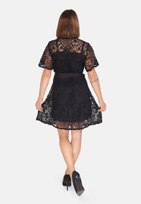 Robe en dentelle noire avec des manches courtes, une taille cintrée et une jupe évasée. Présente des motifs complexes et un ourlet festonné. Portée avec des talons noirs.