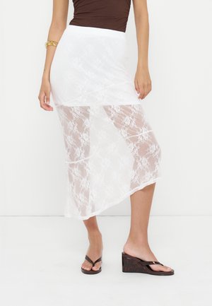 Femme portant une jupe midi en dentelle blanche avec un jupon uni, un haut marron, un bracelet doré et des sandales à talons compensés noires, debout devant un fond blanc.