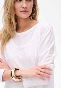 Weißes Langarmshirt mit gerundetem Halsausschnitt, kombiniert mit einem glänzenden goldenen Armreif, der eine glatte Textur und ein einfaches Design zeigt.