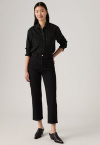 Camicia in denim nero con chiusura a bottoni e due tasche sul petto, abbinata a jeans neri a vita alta. La modella indossa mule nere con tacco a blocco.