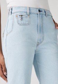 Light blue high-waist jeans with contrast stitching and a small zippered coin pocket, worn with a white top, hand resting on hip.
