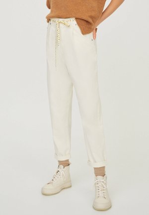 Persona que lleva pantalones blancos de talle alto y corte amplio con cinturón de tela, puños enrollados, calcetines beige, zapatillas blancas altas y un suéter marrón.