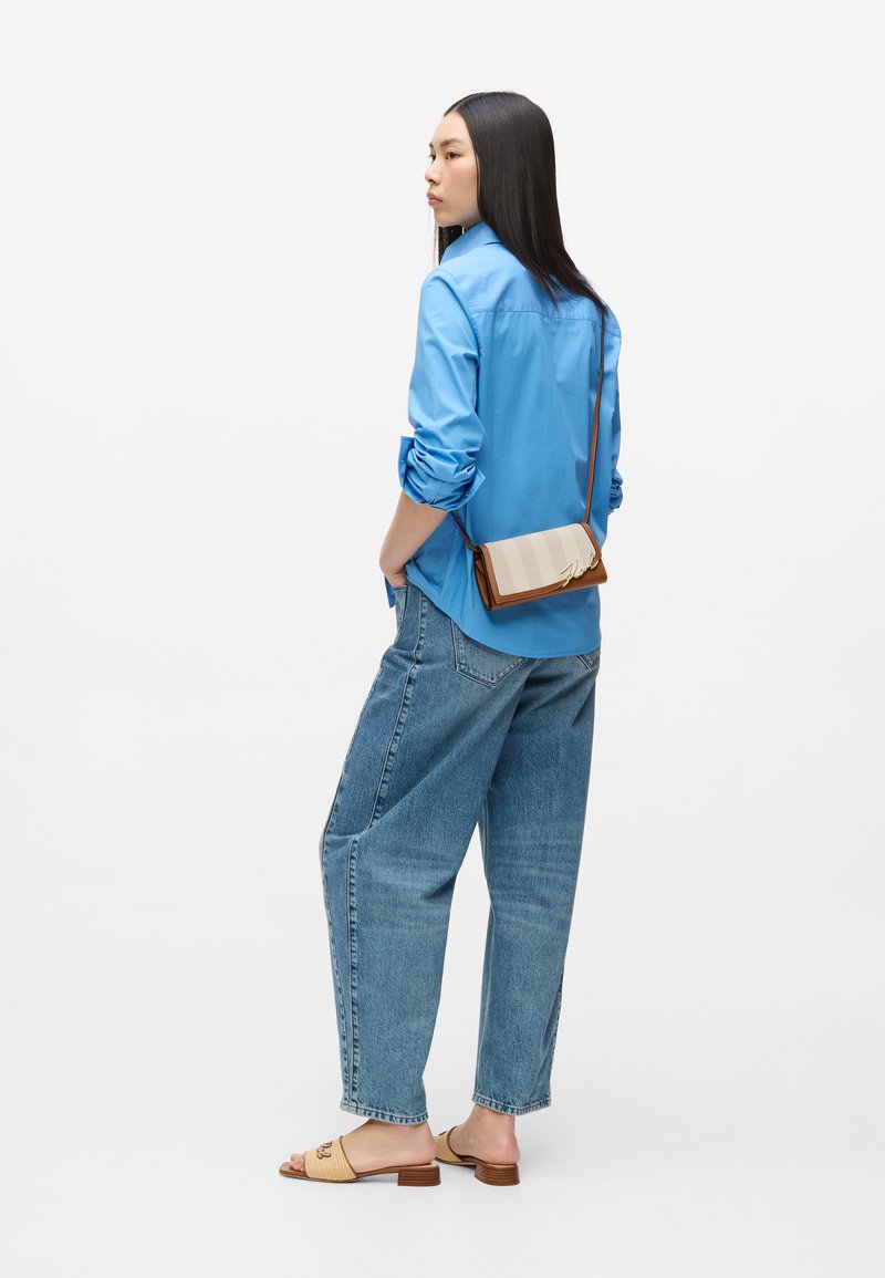 Γυναίκα με μπλε πουκάμισο, φαρδιά τζιν παντελόνια, μπεζ σανδάλια slide και μια μικρή τσάντα χιαστί με ριγέ καπάκι.