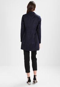 Mørkeblå trenchcoat med en tætsiddende pasform, der har en enkelt bagsøm, båret over sorte tilspidsede bukser og unikke to-farvede hæle.