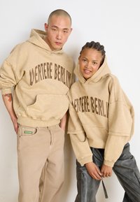 Felpe beige con "VERTERE BERLIN" stampato in marrone scuro. Vestibilità ampia, polsini e orlo a coste, indossate con pantaloni beige e grigi.