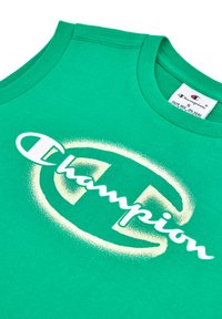 Canotta verde in cotone con un grande design spray bianco riportante la scritta "Champion" e un logo. Tessuto morbido e scollatura arrotondata.