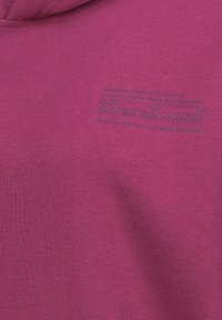 Fuchsiafarbener Kapuzenpullover mit weicher Textur, der entlang der linken Seite einen Schriftzug in kontrastierender Farbe trägt. Einfaches Design, ohne Muster oder Verzierungen.