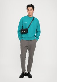 Turquoise sweatshirt with a crew neck, gray tapered pants, and black crossbody bag; black shoes with chunky soles; plain background.