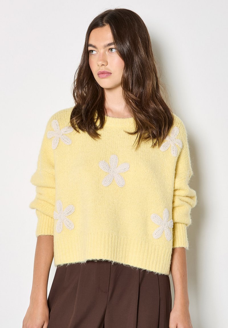 Pull jaune tricoté à coupe ample, orné d'appliqués de fleurs blanches, ourlet côtelé et manches longues. Texture douce et design décontracté.