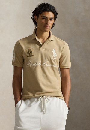 Jeune homme aux cheveux bouclés et foncés portant un polo beige Ralph Lauren et un pantalon blanc à cordon, sur un fond neutre uni.