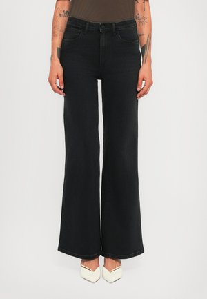 Person wearing high-waisted black wide-leg jeans and white pointed-toe shoes, with visible arm tattoos and no visible top.