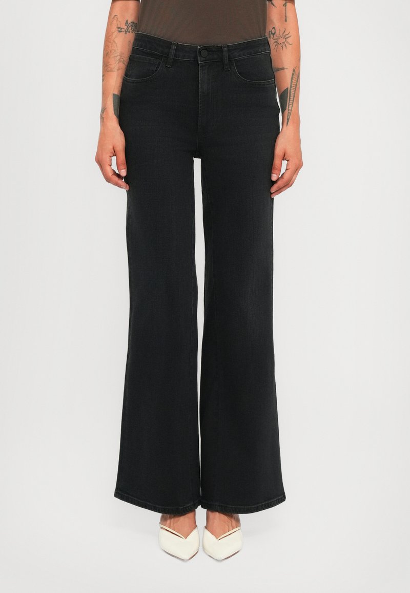 Person wearing high-waisted black wide-leg jeans and white pointed-toe shoes, with visible arm tattoos and no visible top.