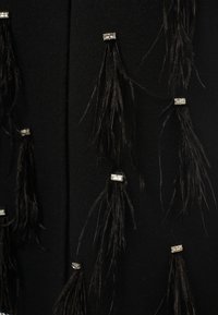 Tessuto nero con lunghe frange nere accentuate da hardware rettangolare trasparente. La texture sembra morbida e opaca.