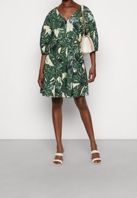 Robe tropicale verte à motif, avec un décolleté en V, des manches bouffantes et une jupe évasée. Assortie à un sac bandoulière crème et des sandales blanches à brides.