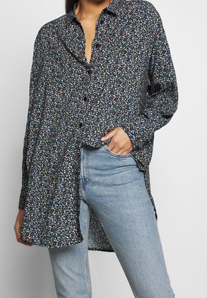 Kobieta ubrana w czarną koszulę z długim rękawem w kolorowy, drobny kwiatowy wzór oraz jasnoniebieskie jeansy, z ręką w kieszeni.