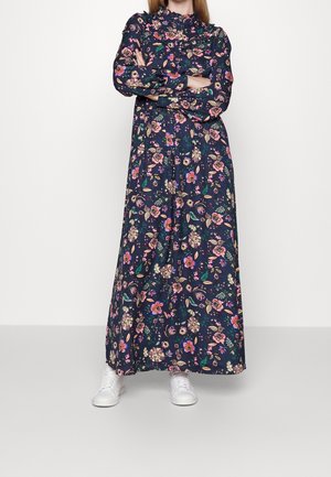 Woman standing with arms crossed wearing a long dark floral dress and white sneakers against a plain light background.