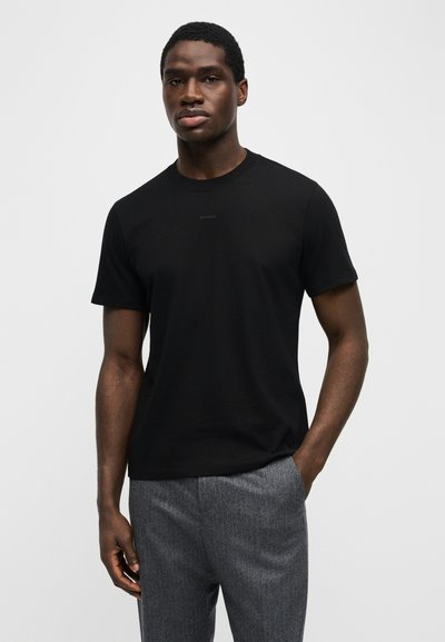 Άνδρας που στέκεται με το ένα χέρι στην τσέπη, φοράει μαύρο μπλουζάκι με κοντά μανίκια και γκρι παντελόνι σε μονόχρωμο φόντο.