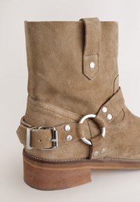 Hellbrauner Wildleder-Stiefelette mit abgerundetem Zehenbereich, silbernen Beschlägen, Schnallendetail und einem Absatz aus Holz. Strukturierte Oberfläche und sichtbare Nähte.