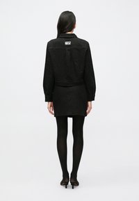 Čierne textúrované sako-šaty s golierom, dlhými rukávmi, zúženým pásom a rovno strihané až po polovicu stehien, v kombinácii s čiernymi pančuchami.