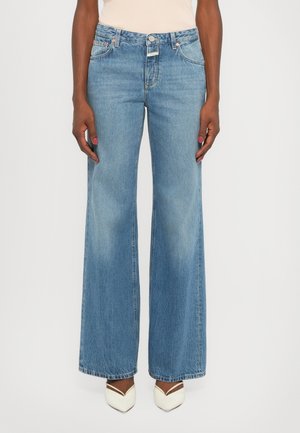 Lys blå vid jeans i denimstoff med blekede detaljer, med fem lommer og en knappelukking. Vist med spisse hvite sko.
