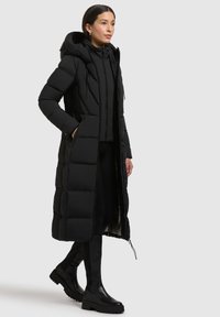 Abrigo puffer negro largo con capucha, que presenta un diseño acolchado, bolsillos laterales y un frente abierto. Llevado con ropa ajustada negra y botas de tobillo.