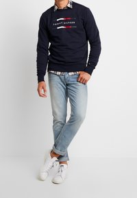 Felpa navy con logo bianco e rosso, indossata sopra una camicia a quadretti, abbinata a jeans azzurri chiari e sneakers bianche. Design casual, vestibilità rilassata.