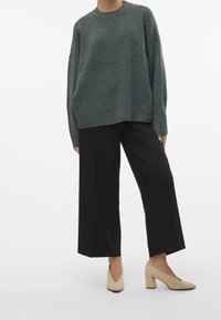 Henkilö, joka pukeutuu tummanvihreään ylisuureen neuleeseen, mustiin leveälahkeisiin lyhyisiin housuihin ja beigekuvioisiin korkokenkiin, seisoo valkoista taustaa vasten.