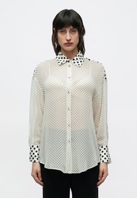 Femeie purtând o bluză albă transparentă cu buline mici negre și guleraș și manșete mari cu buline negre, asortată cu pantaloni negri.