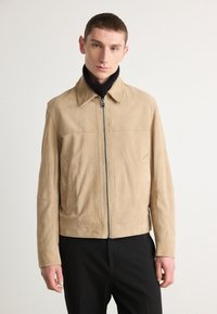 Beige suede jacket with a zip front, pointed collar, two front pockets, and a smooth texture, paired with a black ribbed turtleneck.