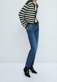 Henkilö, jolla on yllään vihreä-valkoiseraidallinen villatakki, siniset farkut mustalla vyöllä ja mustat kärjistään terävät nilkkasolki kengät, seisoo yksinkertaista seinää vasten.