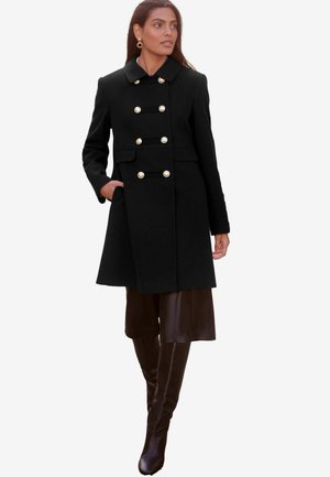 Schwarzer Wollmantel mit goldenen Knöpfen, zweireihiges Design, spitzer Kragen, seitliche Taschen und taillierte Passform. Über einem braunen Outfit getragen.