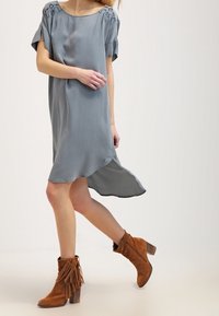 Šedé hedvábné šaty s volnými rukávy, kulatým výstřihem a postranními rozparky, doplněné o hnědé semišové kotníkové boty se třásněmi.