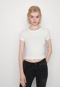 Balta apgriezta T-krekls ar īsām piedurknēm un apaļu kakla izgriezumu, valkāts kopā ar melnajām džinsiem. Gluda auduma, minimālu šuvju un pieguļoša dizaina.