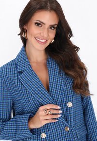 Blazer v modro-turqouise houndstooth vzoru s velkými zlatými knoflíky. Modelka předvádí zlaté kruhové náušnice a dvoubarevný prsten na prstu.