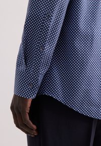 Mano di colore scuro accanto a una manica di camicia blu navy con piccoli pois azzurri e bottoni coordinati, indossata con pantaloni scuri.