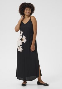 Robe maxi noire sans manches avec imprimé floral d'un côté, présentant des fentes à l'ourlet et une texture lisse et soyeuse. Des ballerines portées avec la robe.