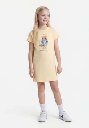 Ung pige i gul kjole med bamseprint, hvide sokker og hvide Nike-sneakers, stående og smilende kiggende til siden.