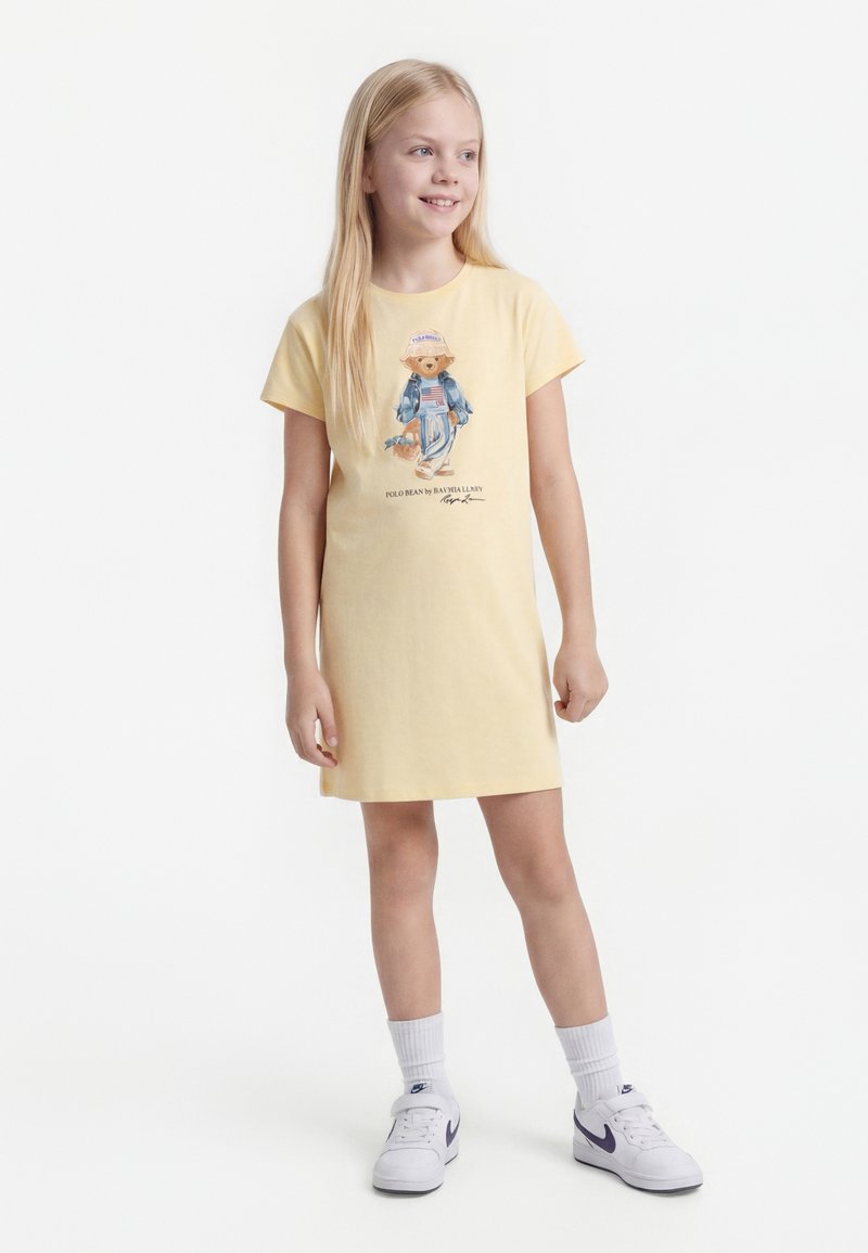 Νεαρή κοπέλα με κίτρινο φόρεμα με γραφικό αρκουδάκι, άσπρες κάλτσες και άσπρα αθλητικά παπούτσια Nike, στέκεται και κοιτάζει πλάγια χαμογελώντας.