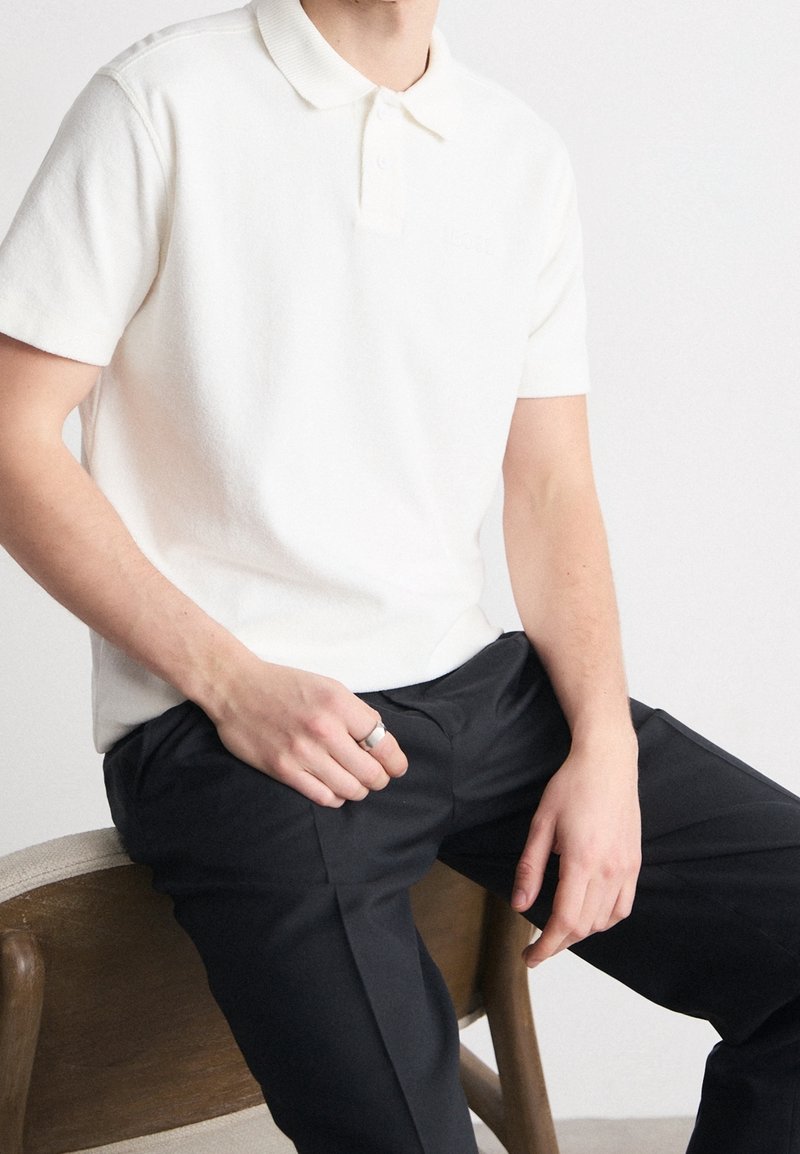 Homme assis sur une chaise en bois, portant un polo blanc à manches courtes et un pantalon noir, avec une bague en argent à la main gauche.