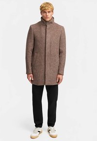 Cappotto marrone in misto lana con collo alto, bottoni frontali e tasche laterali, abbinato a pantaloni neri e sneakers bianche. Design semplice e sartoriale.