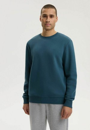 Mężczyzna stojący, ubrany w jednolitą turkusową bluzę z długim rękawem oraz jasnoszare spodnie dresowe, na neutralnym tle.