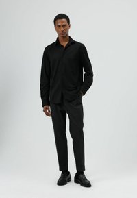 Chemise noire à manches longues avec col, pantalon gris ajusté, et chaussures noires. Texture lisse, coupe ajustée, et lignes épurées. Fond uni.
