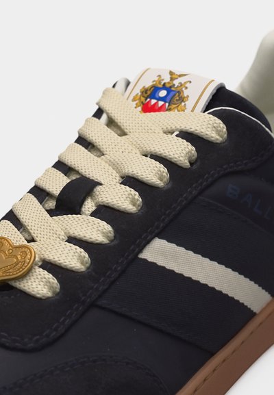 Baskets bleu marine avec lacets crème, rayures latérales blanches, semelle marron et logo en forme de blason sur la languette ainsi qu'une étiquette en métal en forme de cœur sur les lacets.